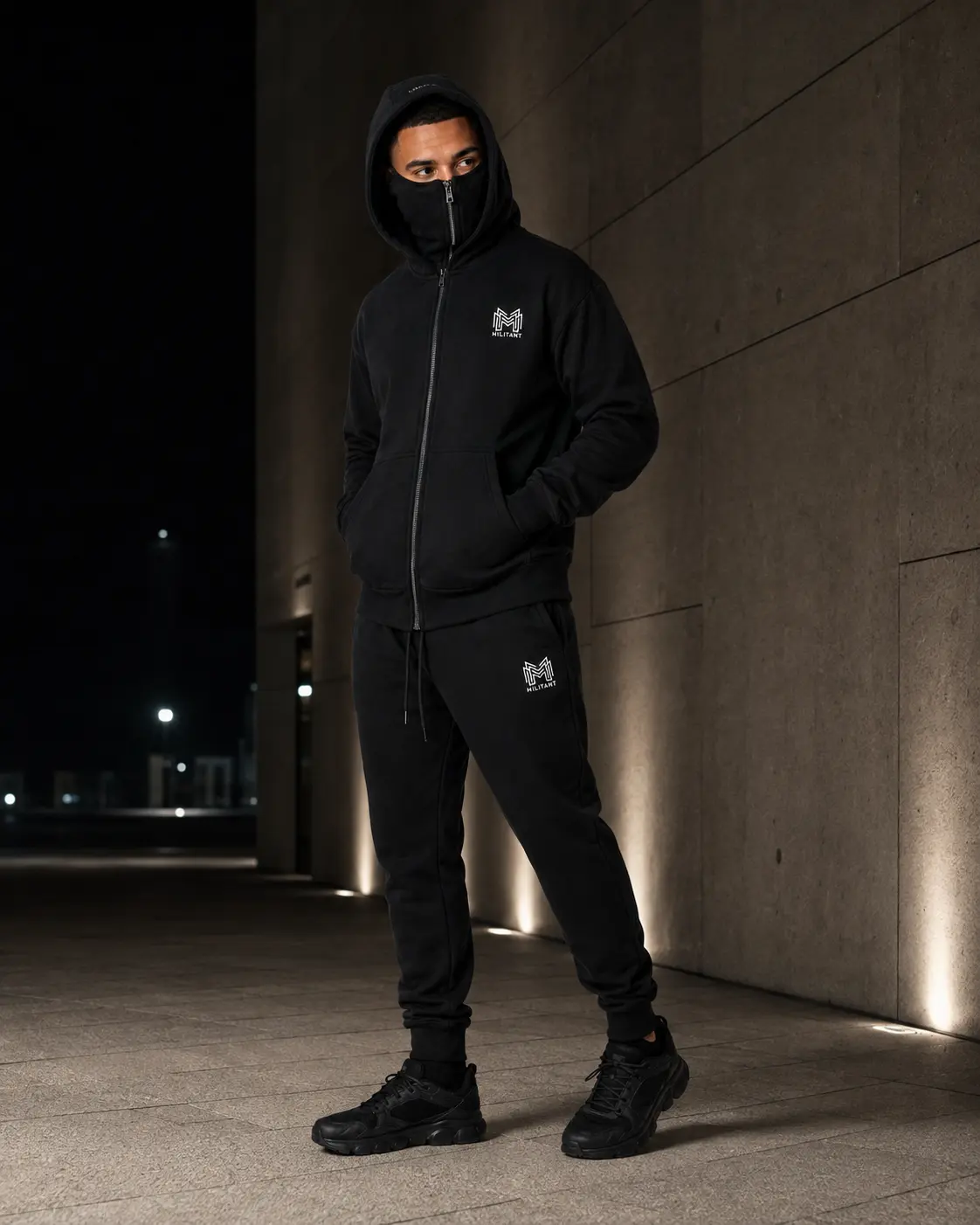 Model wearing the MILITANT tracksuit with hood raised and integrated face cover deployed